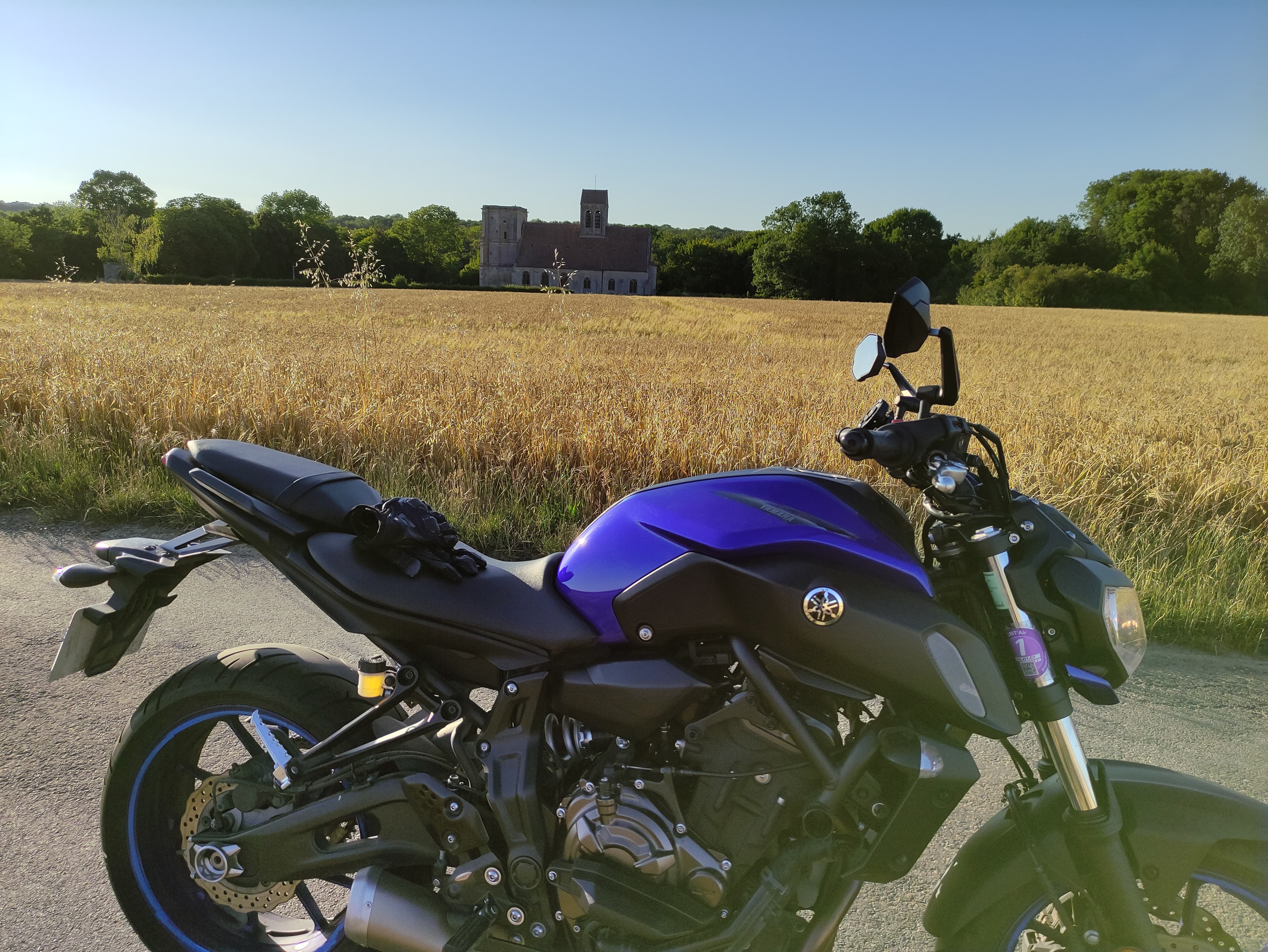 Photo d'une moto devant l'église de Nucourt Une moto bleue au premier plan. Un champ de blé au second plan. L'église de Nucourt dans le Vexin au troisième plan. Le tout sous un ciel bleu dégagé.